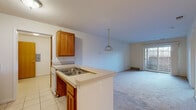 This single-floor unit is designed with a practical, open-concept layout that connects the kitchen and living areas seamlessly. The kitchen is equipped with warm oak cabinetry, a white refrigerator, an electric range, and a double-basin sink set into a peninsula with a light-toned countertop. A pendant light fixture hangs above the peninsula, providing focused illumination. Light tile flooring runs throughout the kitchen, transitioning into plush wall-to-wall carpeting in the living area.

The living space is generously sized and features a sliding glass door that opens to a private outdoor area enclosed by a brick wall, allowing natural light to filter into the room. Vertical blinds provide privacy and light control.

The bedroom is carpeted and includes a closet, with an open doorway connecting to the rest of the unit. The bathroom is finished with light tile flooring and includes accessibility features such as grab bars near the toilet, along with a built-in shelving unit for storage.

A dedicated laundry area houses a full-sized washer and dryer, accompanied by additional built-in shelving for organizational convenience, completing this well-appointed unit.