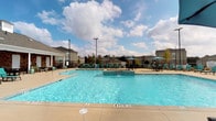 This outdoor amenity space is thoughtfully designed to offer residents a comprehensive recreational experience. At its centerpiece is a large, crystal-clear swimming pool with a raised spa feature, surrounded by a spacious concrete pool deck lined with teal lounge chairs and umbrella-shaded seating areas — ideal for relaxing under open skies.

The pool area is enclosed by a classic black iron fence with brick pillar accents and a secure gated entrance, providing both safety and a polished aesthetic. Lantern-style lighting fixtures mounted on the brick columns add a refined touch to the entryway.

Adjacent to the pool is a generously sized covered outdoor lounge area supported by exposed brick columns. This sheltered patio features a wall-mounted fireplace and television, along with comfortable wicker seating with cushioned upholstery — creating a welcoming space for both casual relaxation and social gatherings. Open-air views of the surrounding community and landscaped grounds extend from this area, enhancing the sense of openness.

A dedicated outdoor grilling station with a gas grill and additional seating rounds out the amenities, offering a convenient space for outdoor dining. Manicured landscaping with mulched planting beds and open green space frame the entire area, contributing to a clean and inviting atmosphere throughout.