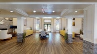 This thoughtfully designed single-floor community facility offers a warm and inviting atmosphere through a cohesive blend of classic architectural details and contemporary furnishings. Rich hardwood flooring flows consistently throughout the entire space, anchoring each area with a sense of warmth and continuity.

The main lounge area serves as the heart of the facility, featuring an open, spacious layout framed by white columns with stacked stone accents. Vibrant yellow sofas and crisp white wingback chairs are arranged to encourage conversation, while large windows dressed in patterned drapery flood the space with natural light and offer views of the surrounding outdoor landscape.

Adjacent to the lounge, a well-appointed dining area accommodates multiple large round and rectangular tables surrounded by upholstered chairs, creating a comfortable and social dining environment. Recessed lighting and additional windows ensure the dining room remains bright and airy throughout the day.

A dedicated seating nook, adorned with a pair of oversized gold-toned art pieces above wainscoted walls, provides a quieter retreat within the larger space. A kitchenette area features granite countertops, a double basin sink, and a beverage refrigerator, supporting the facility's hospitality functions. Decorative artwork with nature-inspired themes is displayed throughout, reinforcing the space's warm and welcoming character.