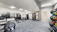 Located in Bixby, Oklahoma, this dedicated fitness center offers a thoughtfully designed exercise space that accommodates a full range of workout needs. The room features a contemporary aesthetic with crisp white walls, elevated ceilings, and stylish patterned carpet flooring in muted blue-grey tones that add visual interest underfoot.

The cardio zone is equipped with multiple treadmills and elliptical machines, arranged to provide ample spacing between users. A large wall-mounted mirror runs along one side of the room, visually expanding the space and allowing exercisers to monitor their form. The strength training area includes a multi-station cable machine, adjustable benches, and a well-stocked dumbbell rack with a range of weights.

A standout design feature is the accent wall adorned with illuminated hexagonal LED light fixtures set against a bold charcoal-grey background. Beneath it, a neatly organized storage rack holds medicine balls, kettlebells, and stability balls in various sizes, making this corner ideal for functional and group training exercises.

Overhead fluorescent strip lighting ensures the entire space is brightly and evenly lit. The room is efficiently arranged to maximize usable floor area while maintaining clear pathways throughout, creating a comfortable and motivating environment for all fitness levels.
