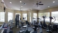 This single-floor fitness center is designed to support a comprehensive workout experience. The space is outfitted with a diverse selection of exercise equipment, including treadmills, stationary bikes, an elliptical, and a multi-station cable weight machine, along with adjustable benches for strength training. The layout provides a comfortable amount of floor space, allowing users to move freely between stations. Neutral-toned walls and a textured ceiling contribute to a clean, functional aesthetic, while crown molding adds a polished architectural touch. Two ceiling fans help maintain airflow and comfort throughout the room during workouts. Multiple large windows line the walls, inviting generous natural light and offering views of the surrounding outdoor greenery, creating a bright and energizing environment. Recessed lighting supplements the natural light, ensuring the space is well-lit at all times. The combination of practical design, varied equipment, and a welcoming atmosphere makes this fitness center a well-rounded facility suited for both cardio and strength-focused training routines.