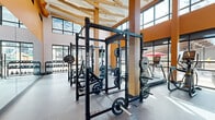 Located at 146 S Wayfinder Ave in Meridian, Idaho, this single-level fitness center is designed to support a wide range of training disciplines in a bright, well-appointed environment. The space is immediately distinguished by its soaring ceilings, exposed timber beam architecture, and warm orange accent walls that lend an energetic yet welcoming atmosphere throughout.

The strength training area is equipped with multiple Rogue power racks loaded with barbells and weight plates, a dedicated dumbbell rack, medicine balls, a plyo box, and a Concept2 rowing machine, providing a comprehensive free weights and functional training zone. A wall-mounted flat-screen TV keeps members informed and entertained during workouts.

The cardio section lines the perimeter with treadmills and elliptical machines, all positioned to take advantage of the floor-to-ceiling windows that flood the space with natural light and offer views of the surrounding outdoor areas, including a glimpsed patio space beyond the glass.

A separate enclosed room, clearly marked as the Fitness Center entrance, features additional cardio equipment such as a stationary bike, along with a large display screen and built-in storage, serving as a versatile studio or supplemental workout space. Rubber flooring throughout ensures durability and safety across all training zones. The open layout maximizes usability while maintaining an airy, spacious feel ideal for focused, high-performance workouts.