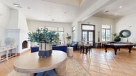 This thoughtfully designed single-story community clubhouse offers a generous floor plan that seamlessly blends social, recreational, and administrative functions. The space is unified throughout by warm terracotta tile flooring, crisp white walls, and dark wood-trimmed doors and windows that lend a cohesive Southwest aesthetic.

The grand entrance greets visitors with soaring ceilings, dramatic dark wood double doors, and a central round wooden table topped with lush botanical décor, setting an inviting tone. Sliding barn doors open to a sunlit lounge featuring a statement white fireplace, plush navy seating, and expansive steel-framed windows that flood the room with natural light and outdoor views.

Adjacent, a casual dining and seating area transitions smoothly into a game room with high ceilings, billiard table, and multiple glass-paneled doors opening to outdoor areas. The kitchen is well-appointed with rich dark wood cabinetry, granite countertops, stainless steel appliances, a large island, and pendant lighting — ideal for community gatherings.

The administrative wing houses multiple private offices furnished with executive desks and leather seating, offering a professional environment with natural light from slatted-blind windows. Together, these spaces create a versatile, welcoming clubhouse that balances comfort, function, and distinct architectural character.