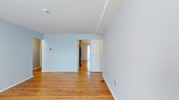This single-floor unit presents a clean and functional layout finished with consistent design details throughout. The living area is characterized by warm-toned hardwood flooring that runs continuously across the space, complementing the soft light blue walls and crisp white trim and baseboards. A window along the exterior wall allows natural light to fill the room, creating a bright and airy atmosphere. A wall-mounted air conditioning unit provides in-unit climate control, accompanied by baseboard heating along the perimeter walls, ensuring year-round comfort. The entry door is positioned along one wall, offering a straightforward and accessible entry point into the space. A structural beam is visible along the ceiling, adding subtle architectural definition between areas. An open doorway visible from the main living space leads to an adjacent room, where a stainless steel appliance is partially visible, suggesting a kitchen or utility area nearby. The unit's overall aesthetic is minimal and neutral, making it highly adaptable to a variety of furnishing styles. The continuous hardwood flooring, neutral palette, and practical amenities come together to form a cohesive, move-in-ready living environment.