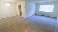 This single-floor residence offers a clean, functional layout designed for comfortable everyday living. The main living area is generously sized, finished with neutral-toned wall-to-wall carpeting that adds warmth underfoot. A large window along one wall allows natural light to fill the room, creating a bright and airy atmosphere. A baseboard heating unit is positioned beneath the window, ensuring year-round comfort.

The open layout connects the living area directly to the kitchen, which is accessible through a doorway framed by bi-fold or standard doors. The kitchen features warm wood-toned cabinetry that provides ample storage, along with countertop space suitable for meal preparation. Appliances are visible within the kitchen, suggesting a practical and ready-to-use setup.

The walls throughout are painted in a neutral off-white tone, offering a versatile backdrop that complements a variety of furnishing styles. A thermostat is mounted on the wall near the kitchen entry, allowing easy climate control. Overall, the space presents a straightforward, well-maintained interior with a functional flow between the living and kitchen areas.