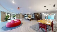 Located at 113 Ellington Blvd in Gaithersburg, Maryland, this expansive single-floor community lounge offers a generously scaled, open-plan layout thoughtfully divided into several distinct functional zones. The entrance area greets visitors with large floor-to-ceiling glass doors framed by tall indoor plants, flooding the space with natural light and offering clear views to the surrounding streetscape. Light-toned large-format tile flooring anchors the entry and kitchen areas, transitioning to patterned carpet throughout the lounge zones.

A bold circular red sofa serves as a striking centerpiece near the entry, setting an energetic, contemporary tone. Adjacent to it, a kitchen bar area features a dark-finished island with bar seating, pendant lighting, and a decorative mosaic tile accent wall. A built-in dark wood cabinetry unit with an integrated flat-screen TV provides a sophisticated media focal point.

The lounge extends into multiple relaxed seating areas, each thoughtfully arranged with upholstered sofas, accent chairs, and coffee tables in a neutral palette punctuated by vibrant yellow, red, and navy accents. Curated wall art, lush potted plants, and draped curtain dividers add warmth and definition to the space. A built-in workstation with monitors offers a practical workspace. Recessed lighting throughout ensures a bright, welcoming ambiance across the entire floor.
