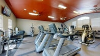 This fitness center offers a comprehensive workout environment designed to support a wide range of exercise routines. The space is immediately distinguished by its striking deep-red ceiling, which adds a bold, energizing aesthetic contrast to the neutral-toned walls. Ceiling fans are positioned throughout to promote air circulation, complementing the recessed and flush-mounted lighting that keeps the space bright and functional.

The cardio zone is well-stocked with a row of Matrix treadmills and Matrix elliptical trainers, providing ample options for aerobic exercise. Wall-mounted flat-screen televisions are positioned for easy viewing from the cardio equipment. A dedicated strength and free weights area features a multi-rack dumbbell station with an extensive weight selection, a weight bench, and a multi-functional cable machine for resistance training. Additional strength equipment is arranged throughout, including a leg press and upper-body machines.

Large windows fitted with blinds allow natural light to filter into the space, enhancing the open, airy feel. Durable rubber flooring covers the workout areas, offering both safety and comfort. Full-length mirrors along one wall expand the sense of space visually while allowing users to monitor their form. The layout is thoughtfully organized, separating cardio from strength training zones for an efficient, distraction-free experience.