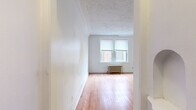 This single-floor unit offers a straightforward, functional layout across several distinct rooms, all finished with warm hardwood flooring throughout. The living area is notably open and bright, featuring dual windows that allow natural light to fill the space. A cast-iron radiator beneath the windows provides classic steam heating, a characteristic detail repeated across multiple rooms in the unit. A textured ceiling adds architectural character overhead, while crisp white walls keep the overall aesthetic clean and neutral.

The layout connects the living area to an additional room, also finished with hardwood floors and its own set of windows overlooking neighboring brick buildings. This secondary space is versatile and well-proportioned for use as a bedroom or additional living area.

The kitchen is a compact, efficiently arranged space featuring white cabinetry, wood-trimmed countertops, a stainless steel sink, and a full-size refrigerator. Natural light enters through a side window, and wood-look flooring ties the space together visually.

The bathroom is finished in an all-white palette with wood-look flooring, a full soaking tub with an overhead window, a single vanity with storage, and a wall-mounted mirror. A radiator provides warmth, consistent with the building's heating system throughout.