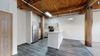 Located at 550 W Western Ave in Muskegon, Michigan, this single-floor loft-style unit masterfully blends historic industrial character with contemporary updates. The open-concept living and kitchen area is anchored by soaring wood-plank ceilings supported by heavy timber beams and original structural posts, creating a warm, raw aesthetic that defines the space. Exposed ductwork and pendant industrial lighting complement the building's heritage.

The kitchen is outfitted with modern light-toned cabinetry, white countertops, and a full suite of stainless steel appliances — including a refrigerator, electric range, and over-the-range microwave — all set atop durable wood-look plank flooring. A peninsula provides additional workspace and casual seating.

The bedroom showcases the building's most striking original details: a full exposed stone masonry wall alongside a section of original brick, framed by low-profile windows that allow natural light to filter in. Carpeting adds comfort underfoot, while the timber ceiling and visible HVAC ducts maintain the industrial character throughout.

The bathroom is clean and functional, featuring a full tub-shower combination, modern vanity with wood-toned cabinetry, and wood-look flooring that ties the unit together cohesively. This space offers a compelling combination of authentic historic texture and thoughtfully updated living amenities.