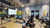This well-appointed fitness studio is designed to support a wide range of workout routines in a clean, contemporary environment. The space is anchored by a row of Matrix stationary spin bikes arranged on durable rubberized flooring with blue accent mats, providing a dedicated cycling zone for high-energy cardio sessions. Striking floor-to-ceiling mirror panels framed in sleek black steel line the walls, visually expanding the space and allowing users to monitor their form throughout their workouts. A large flat-screen display is mounted within the mirrored wall, ideal for streaming guided fitness classes or entertainment during sessions. The studio also features a well-stocked equipment rack holding an assortment of colorful stability balls, medicine balls, and foam rollers, making it fully equipped for functional and group fitness training. Soft blue-grey walls are accented by framed abstract artwork, lending a polished, upscale aesthetic to the room. Recessed lighting throughout ensures the space is consistently well-lit, while a coffered ceiling detail adds an architectural touch. An open glass partition connects the studio to an adjacent fitness area, creating a seamless and expansive wellness environment overall.