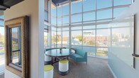 This single-floor space offers a compelling blend of modern design and functional living. A standout feature is the expansive lounge or common area enclosed by towering floor-to-ceiling glass curtain walls, allowing an abundance of natural light to flood the interior while providing sweeping views of the surrounding urban landscape. This bright, open seating nook is furnished with a round table, teal sofa, and colorful accent stools, creating a welcoming collaborative environment. The remaining living areas transition seamlessly, featuring rich wood-style plank flooring in the main living and entry zones, which flows into plush carpeting in additional rooms. Crown molding and clean architectural columns add refined detail throughout. The kitchen area is highlighted by elegant granite countertops and pendant lighting, contributing a polished aesthetic. Neutral, warm-toned walls throughout create a cohesive and inviting ambiance. Doorways connect to additional rooms, suggesting a well-organized and functional layout. The entry features a paneled white door with a keypad access system, emphasizing both security and convenience. Overall, the space combines contemporary architectural elements with practical design for a comfortable and stylish environment.