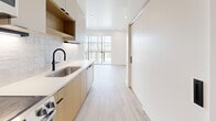 Located at 634 S 400 W in Salt Lake City, Utah, this modern studio offers a thoughtfully designed single-floor layout that maximizes every square foot. The open-concept living, dining, and kitchen area is anchored by light-toned luxury vinyl plank flooring that flows seamlessly throughout, complemented by crisp white walls and recessed lighting that keep the space feeling bright and airy.

The galley-style kitchen is a standout feature, outfitted with two-tone cabinetry pairing warm natural wood tones with soft off-white uppers. Quartz countertops, a textured tile backsplash, a matte black faucet, and stainless steel appliances — including a range, dishwasher, microwave, and full-size refrigerator — deliver a polished, functional cooking space. A large window at the end of the kitchen corridor draws in natural light and frames views of the surrounding cityscape.

The open living area is generously proportioned, with clean sightlines and neutral finishes that offer a versatile canvas for furnishing. Two closets provide ample storage.

The bathroom continues the modern aesthetic with light wood vanity cabinetry, a white quartz countertop, a matte black faucet, an arched black-framed mirror, and full-height vertical tile surrounding a soaking tub and shower combo. Built-in open shelving adds practical storage. The overall ambiance throughout is contemporary, clean, and move-in ready.
