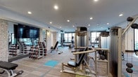 This fitness center offers a comprehensive workout environment designed to accommodate a variety of exercise routines. The space features a thoughtfully arranged layout with distinct zones for both strength training and cardiovascular exercise. A substantial free weight section lines one wall, equipped with a full rack of dumbbells alongside adjustable weight benches. Multiple cable and resistance machines from Precor are positioned throughout the floor, providing options for full-body strength training. Cardio equipment, including ellipticals and upright machines, is situated near the windows, allowing users to exercise with the benefit of natural light. The ceiling is fitted with recessed lighting that ensures consistent illumination throughout the space. A large flat-screen television is mounted on the wall, adding an entertainment element to workouts. The flooring consists of carpet tiles with accents of blue, providing both durability and cushioning underfoot. Stone-accented columns add a decorative architectural touch, contributing to the modern and polished aesthetic of the facility. Large windows along the perimeter allow natural light to fill the room, creating an open and energizing atmosphere.