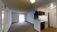 This single-floor space showcases an open-concept design that seamlessly connects the kitchen and living area. The kitchen features dark cabinetry that provides a striking contrast against the light-colored walls, and includes a white electric range and countertop space along a peninsula that partially separates it from the main living area. Under-cabinet lighting adds functional illumination to the workspace. The living area is finished with wall-to-wall carpeting, contributing to a warm and comfortable atmosphere. A sliding glass door dressed with vertical blinds anchors one wall, allowing natural light to fill the space and offering potential access to an outdoor area. A pendant light fixture is visible overhead, providing ambient lighting. The textured ceiling adds a subtle architectural detail throughout. The overall color palette is neutral, with beige and white tones dominating the space, creating a clean and versatile backdrop. The open layout and connected design of the kitchen and living areas make the space feel functional and adaptable for everyday living.