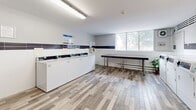 Located at 86 Cedar St S in Kitchener, Ontario, this shared laundry facility offers a clean, functional, and well-maintained space designed to meet residents' everyday laundry needs. The room is finished with light wood-look luxury vinyl plank flooring and white walls accented by a horizontal band of dark tile, giving the space a modern and tidy aesthetic. Bright fluorescent lighting ensures excellent visibility throughout.

Along one wall, a row of commercial-grade front-load washing machines and dryers lines the space, each equipped with digital controls for ease of use. Instructional signage is posted above the machines for user guidance. A centrally positioned large folding table with a stone-look surface and stainless steel legs provides ample space for sorting and folding laundry. A utility sink offers additional convenience for hand-washing or pre-treating garments.

Natural light filters in through a set of windows overlooking a tree-lined outdoor area, adding a welcoming brightness to the room. A decorative potted plant adds a touch of greenery to the otherwise utilitarian environment. The space is accessed through a secure teal-painted door and is equipped with a smoke detector and fire safety equipment, ensuring a safe environment for all users.