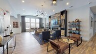 This single-floor community amenity center in Lockport, Illinois, is thoughtfully designed to offer residents a variety of social and recreational spaces. The centerpiece of the space is a spacious lounge area anchored by a striking floor-to-ceiling feature wall with reclaimed wood-style tile, a linear fireplace, and built-in shelving adorned with natural log accents. A wall-mounted TV completes the entertainment setup, while an eclectic sputnik chandelier and ceiling fans illuminate multiple seating arrangements, including leather armchairs, upholstered sofas, and accent chairs arranged on patterned area rugs. Warm hardwood-style flooring flows throughout the entire space. Large windows along the perimeter flood the lounge with natural light and offer views of the surrounding community. Adjacent to the lounge is a well-appointed kitchen featuring white cabinetry, quartz countertops, a decorative tile backsplash, stainless steel appliances, and a central island with leather barstools beneath elegant pendant lighting. A casual dining nook with tables and chairs sits nearby, bathed in natural light from a bank of windows. The game room, visible from the lounge, provides additional recreational space. A fully equipped fitness center rounds out the amenities, housing a wide range of cardio and strength training equipment.
