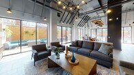 Located at 55 S Vail Ave in Arlington Heights, Illinois, this single-floor amenity space combines industrial-modern design with comfortable, functional gathering areas. The expansive open-plan layout features soaring ceilings with exposed steel beams, dark structural elements, and a network of pendant lighting that creates a warm, inviting atmosphere throughout.

The main lounge area is anchored by a sleek fireplace with a wall-mounted television above, flanked by plush sectional seating and accent chairs arranged on area rugs over large-format tile flooring. A built-in bar counter with stools provides an additional casual seating option. Floor-to-ceiling sliding glass doors line the exterior walls, flooding the space with natural light and offering direct views and access to the outdoor pool and patio area.

The kitchen and dining zone is appointed with stainless steel appliances, including a full-size refrigerator and microwave, a double-basin sink, white quartz countertops, warm wood cabinetry, and a striking deep-blue chevron-patterned tile backsplash with open floating shelves. A large communal wood-topped dining table with industrial-style metal legs and leather bar stools serves as the centerpiece of this area.

The outdoor pool area, visible through the expansive glass doors, features a full-size swimming pool surrounded by a generous patio with lounge seating.
