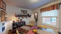 Located at 900 Hill St in Greensboro, NC, this single-story home offers a warm, eclectic aesthetic defined by carefully curated vintage and antique elements throughout. The living area serves as the heart of the space, featuring carpeted flooring layered with patterned area rugs, a large rustic armoire with ornate detailing, and an assortment of comfortable seating. Windows dressed with fabric valances allow natural light to filter in, enhancing the cozy, lived-in atmosphere. A bookshelf near the entry door displays collectibles and framed artwork, contributing to the home's richly personalized character.

The dining area continues the vintage theme with a rustic wooden table, yellow accent chairs, and a wall-mounted shelf displaying pitchers, hats, and framed prints. An open shelving unit holds an assortment of pottery and dishware, adding functional charm to the space.

The bedroom features warm hardwood flooring, a classic brass bed frame, a patchwork quilt, and a Tiffany-style lamp, evoking a nostalgic, timeless feel. Multiple windows provide ample natural light.

The kitchen is compact and functional, equipped with white cabinetry, a coil-burner range, and a double sink with butcher block countertops. A door with a window leads to the exterior. The bathroom is clean and simply appointed, with a shower/tub combination and a decorative vintage street sign as a distinctive accent.