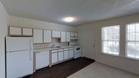 This single-floor space features an open kitchen and living area designed for practical, everyday use. The kitchen is outfitted with white cabinetry accented by light wood trim, offering generous upper and lower storage. A full suite of white appliances includes a refrigerator, electric range with overhead exhaust, and dishwasher. The countertops are light-toned, complementing the overall neutral color palette, while a stainless single-basin sink sits beneath a window that allows natural light to enter. The kitchen floor features dark hardwood-style flooring that provides a strong contrast to the white cabinetry and appliances. A door with an adjacent window on the far wall provides additional natural light and likely serves as a secondary entry point. The living area transitions from the hard flooring to carpet, creating a distinct and comfortable zone. A textured ceiling with a flush-mount light fixture provides ambient illumination throughout. The overall design is clean and straightforward, emphasizing functionality with a cohesive, understated aesthetic.