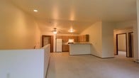 This single-floor space offers a practical and welcoming open-concept layout that seamlessly connects the living area and kitchen. The living area is finished with plush wall-to-wall carpeting in a neutral tone, providing a comfortable and cohesive foundation for the space. Recessed lighting and a ceiling fan overhead ensure the area remains well-lit and properly ventilated.

The kitchen is positioned at the back of the open floor plan and features warm honey-toned wood cabinetry that lines both the walls and base, offering ample storage. White appliances, including a refrigerator and range with overhead ventilation, are neatly integrated into the layout. A raised breakfast bar peninsula separates the kitchen from the living area, creating a natural transition point that is ideal for casual dining or entertaining.

The flooring in the kitchen transitions to a smooth, easy-to-clean surface, contrasting with the carpeted living space. The walls throughout are finished in a clean, neutral tone that enhances the sense of openness. A doorway visible in the background hints at additional rooms beyond. Recessed and flush-mount ceiling fixtures provide consistent illumination throughout, contributing to the bright and functional ambiance of this straightforward, well-organized space.