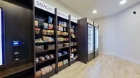 Located at 870 S SH 161 Frontage Rd in Grand Prairie, Texas, this neatly appointed convenience market offers a thoughtfully designed retail experience within a compact, single-room layout. The space features light hardwood-style flooring that contrasts cleanly with crisp white walls and recessed ceiling lighting, creating a bright and welcoming atmosphere.

Along one wall, a series of tall, dark-stained wood shelving units are organized into clearly labeled sections — "Snacks," "Refresh," "Enjoy," and "Refuel" — providing an intuitive shopping experience. The shelves are fully stocked with a wide variety of packaged snacks, dry goods, and sundry items, with woven basket organizers adding a touch of visual warmth. Adjacent to the shelving, glass-door refrigeration units offer chilled and frozen product selections.

A small, built-in storage cabinet sits near the entry, serving as a checkout or service station. The space is accessed through striking floor-to-ceiling black-framed glass sliding doors, which provide a view into an adjoining fitness facility, suggesting this market serves as a convenient amenity within a larger residential or hospitality complex. The overall design is modern and clean, balancing functionality with an upscale aesthetic.