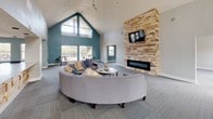 This single-floor community clubhouse is defined by its striking architectural design and open, welcoming atmosphere. The centerpiece of the space is a dramatic vaulted ceiling that soars to impressive heights, accentuated by a bold teal accent wall and a wall of clerestory windows that flood the interior with natural light. A floor-to-ceiling stacked stone fireplace serves as a commanding focal point in the main lounge area, complemented by a mounted flat-screen TV above a sleek linear firebox. The lounge is furnished with a generously sized curved sectional sofa, creating an inviting gathering space.

Patterned carpet tiles in soft gray tones run throughout the main areas, lending a clean, contemporary feel. Multiple seating zones are thoughtfully arranged throughout the space, including wingback leather chairs and stylish accent furniture. French doors open to an outdoor patio, while a series of windows along the teal-accented walls bring in views of the surrounding neighborhood.

The full kitchen features dark cabinetry, stainless steel appliances, open shelving, and an expansive quartz-topped island with a built-in sink — ideal for events and gatherings. Ceiling fans are installed throughout for added comfort. The overall ambiance balances modern design with a warm, communal character.