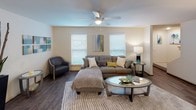 This two-floor residence offers a thoughtfully designed layout with a cohesive, neutral color palette and wood-look flooring running throughout the main living areas. The living room is a comfortable, well-lit space featuring multiple windows with slatted blinds that invite natural light, complemented by a ceiling fan overhead and warm hardwood-style flooring underfoot.

The kitchen follows a galley configuration, lined with light wood cabinetry, granite-style countertops, and black appliances including a range and refrigerator. A pantry closet at the end of the kitchen provides additional storage space.

The space includes two bedrooms, each with ceiling fans, carpeted floors, and windows that bring in natural light. Both rooms offer ample space for furnishings and feature neutral wall tones that lend a calm, restful ambiance.

The bathrooms are finished consistently with granite-style countertops, wood-tone cabinetry, and dark hardwood-style flooring. The primary bathroom includes a wide vanity with an extended mirror and a tall storage cabinet, while a secondary bathroom features a full tub and shower enclosure with white tile surround. A compact water closet serves as an additional convenience on the upper floor.

Overall, the space presents a clean, functional, and welcoming environment with consistent design elements carried throughout every room.
