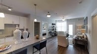This single-story residence at 4625 Fairview Dr in Wilmington, North Carolina, showcases a thoughtfully designed open-concept floor plan that connects the kitchen, dining, and living areas into one cohesive, functional space. Dark hardwood flooring runs throughout, providing a rich contrast against the light gray walls and white trim that define the interior palette.

The kitchen features white cabinetry, a granite-topped island with bar seating, and pendant lighting that adds a stylish focal point overhead. The open layout allows the kitchen to flow naturally into the living area, which is bright and welcoming, benefiting from large windows that invite natural light and offer views of the outdoors. A ceiling fan with light fixture serves the living space, enhancing comfort and airflow.

Frosted glass interior doors provide a contemporary touch while maintaining privacy between rooms. The bedroom, visible from the main living area, continues the same refined aesthetic with coordinated furnishings and ample light. The overall ambiance is modern and clean, with a neutral color scheme and quality finishes that create a polished, move-in-ready environment.