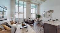 Located in the heart of Columbus, Ohio, this two-story urban loft on East Spring Street masterfully blends industrial character with contemporary finishes. The main level opens through a welcoming hallway with decorative slatted ceiling details and wood-look flooring that continues throughout. The living and dining area is an immediate showstopper, boasting soaring double-height ceilings and an expansive wall of floor-to-ceiling grid windows that floods the space with natural light and provides direct access to an outdoor balcony. The open-concept layout flows seamlessly into the dining area and kitchen, where dark shaker-style cabinetry, granite countertops, a subway tile backsplash, a large island with bar seating, and a full suite of stainless steel appliances create a highly functional and stylish cooking space. A clean, neutral bathroom with granite counters and terrazzo-style flooring completes the main level. The second floor houses a generously sized bedroom with plush carpet, exposed ductwork adding to the loft aesthetic, and an open overlook to the living area below. A spacious walk-in closet with built-in shelving and wood flooring provides ample storage. This loft offers a distinctive urban living experience in a prime downtown Columbus location.