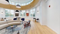 Located at 6515 Glen Erin Dr in Mississauga, Ontario, this architecturally distinctive single-floor living space makes an immediate impression with its bold circular layout and soaring double-height ceilings. A continuous band of warm natural wood wraps the upper interior curve, creating a striking contrast against the crisp white walls below and lending the space a refined, modern character.

Light pours in generously through a wrap-around arrangement of large windows positioned at both the upper and lower levels, offering views of the surrounding greenery and filling the room with a bright, airy atmosphere throughout the day. Light-toned hardwood flooring runs throughout, further enhancing the sense of openness and warmth.

The living area is thoughtfully arranged to balance both social and intimate settings. At the center, a sculptural curved sofa anchors a generous seating area defined by a layered area rug, complemented by mid-century-inspired lounge chairs in grey and warm wood tones. Surrounding the perimeter, compact bistro tables paired with terracotta-hued upholstered chairs provide inviting spots for smaller gatherings or quiet moments.

Carefully selected artwork adorns the white walls, adding pops of color and artistic depth. Statement pendant lighting hangs from above, contributing both function and visual interest. The overall ambiance is one of sophisticated comfort — a well-designed space that balances contemporary elegance with an inviting, community-oriented warmth.