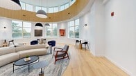 Located at 6515 Glen Erin Dr in Mississauga, Ontario, this architecturally distinctive single-floor living space makes an immediate impression with its bold circular layout and soaring double-height ceilings. A continuous band of warm natural wood wraps the upper interior curve, creating a striking contrast against the crisp white walls below and lending the space a refined, modern character.

Light pours in generously through a wrap-around arrangement of large windows positioned at both the upper and lower levels, offering views of the surrounding greenery and filling the room with a bright, airy atmosphere throughout the day. Light-toned hardwood flooring runs throughout, further enhancing the sense of openness and warmth.

The living area is thoughtfully arranged to balance both social and intimate settings. At the center, a sculptural curved sofa anchors a generous seating area defined by a layered area rug, complemented by mid-century-inspired lounge chairs in grey and warm wood tones. Surrounding the perimeter, compact bistro tables paired with terracotta-hued upholstered chairs provide inviting spots for smaller gatherings or quiet moments.

Carefully selected artwork adorns the white walls, adding pops of color and artistic depth. Statement pendant lighting hangs from above, contributing both function and visual interest. The overall ambiance is one of sophisticated comfort — a well-designed space that balances contemporary elegance with an inviting, community-oriented warmth.