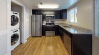 This single-floor residence offers a well-organized layout with distinct living, sleeping, and functional areas. The open living and dining area is finished with warm hardwood flooring and neutral walls, allowing ample natural light to flow in through windows fitted with blinds. Track lighting accents the space, adding a modern touch to the otherwise clean aesthetic.

The kitchen is efficiently designed with dark espresso cabinetry that contrasts sharply with granite countertops. Stainless steel appliances — including a refrigerator, range, microwave, and dishwasher — provide a cohesive, contemporary look. Light hardwood flooring runs throughout this area, contributing to a bright and functional workspace.

The bedroom features plush wall-to-wall carpeting and neutral beige tones, with two windows that invite natural light and views of surrounding greenery. The room provides a calm, comfortable atmosphere.

The bathroom is appointed with a geometric black-and-white tile floor, a dark-cabineted vanity topped with granite, and an oversized mirror. A tiled tub-and-shower combination occupies one end, with a window adding natural light. In-unit stacked laundry is also present, adding practical convenience to the overall layout.