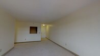 This single-floor space presents a straightforward, open layout finished in neutral tones throughout. The main living area is carpeted and benefits from smooth, white ceilings with flush-mount lighting that evenly illuminates the room. The walls are painted in a clean off-white, contributing to a bright and airy atmosphere. A notable architectural feature is the pass-through window cutout in the partial wall, which connects the living area to an adjacent space, likely a kitchen. This detail adds a degree of openness and allows light to flow between areas. A white door visible in the background leads to an entryway or hallway, which appears to transition into additional rooms or an exit. The space is compact yet functional, with a simple, efficient design. Electrical outlets are positioned along the baseboards, and the clean lines of the room suggest a well-maintained interior. Overall, the space offers a neutral, move-in-ready environment suitable for a variety of furnishing arrangements and uses.