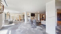 This elegantly designed single-floor amenity space offers a seamless blend of luxury and functionality across a series of distinct yet interconnected areas. The main lounge is anchored by a marble-clad fireplace wall with a mounted flat-screen TV, surrounded by stylish woven lounge chairs set atop a geometric-patterned carpet in shades of grey and blue. Floor-to-ceiling glass partitions allow natural light to flow throughout while maintaining visual continuity between zones.

A dedicated music room features a grand piano as its centerpiece, complemented by a metallic gold ceiling, a sculptural pendant light, and bold abstract artwork, creating an intimate and dramatic ambiance. Adjacent social areas include a wet bar with pendant lighting, bar seating, a mounted display screen, and a textured accent ceiling — ideal for casual gatherings.

The library lounge showcases geometric honeycomb shelving in warm gold tones and mirrored display units, flanked by a striking graphic feature wall in black and white. A bright seating area with floor-to-ceiling windows offers lush outdoor views and abundant natural light. Private study nooks and a restroom round out the practical amenities. Light hardwood flooring and patterned carpeting unify the space with a refined, contemporary aesthetic throughout.