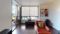 This well-appointed single-floor residence offers a thoughtfully designed open-concept layout that balances modern aesthetics with comfortable living. The living area is bright and inviting, featuring rich hardwood flooring, large floor-to-ceiling sliding glass doors that open to a balcony, and views of surrounding palm trees and the urban skyline. Roller shades provide light control while maintaining the airy feel of the space. Decorative sunburst mirrors and colorful accent pillows add a curated, contemporary touch to the lounge area. The kitchen is efficiently laid out with sleek dark cabinetry, white quartz countertops, a stainless steel sink, and a full suite of stainless steel appliances including a refrigerator, microwave, and range. An accent wall in muted blue adds a distinctive design element that carries into the secondary bedroom, which also features a built-in open shelf unit and direct access to a closet and bathroom. The primary bedroom is serene and spacious, with plush carpeting, symmetrical nightstands, round mirrors, and soft neutral tones that create a restful atmosphere. The bathroom is clean and modern, showcasing dark wood vanity cabinetry, a white quartz countertop, a large wall mirror, and a glass-enclosed shower-tub combination with tiled walls.