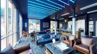 The Community Room at 28 Southfield Ave in Stamford, Connecticut is a thoughtfully designed shared space that balances vibrant aesthetics with practical amenities. The room's most striking feature is its ceiling installation of parallel cyan neon light strips suspended against a deep navy backdrop, creating a dramatic and modern atmosphere throughout. Deep blue walls carry the color theme consistently, complemented by richly patterned blue carpeting that evokes a nautical sensibility. The lounge area offers generous seating through a combination of navy blue sofas, tufted leather armchairs in warm cognac brown, and coffee tables styled with curated décor accents. Floor-to-ceiling glass windows and sliding doors line one wall, flooding the space with natural light and providing open views of the surrounding neighborhood. A dedicated bar-height seating section with sleek stools offers an additional social gathering spot. The fully equipped kitchen features white cabinetry, stainless steel appliances including a refrigerator and wall ovens, and a gold-accented island with bar seating — ideal for hosting events. Decorative pendant lighting in geometric gold fixtures adds warmth and contrast to the cool blue palette. Framed artwork and nautical-inspired decorative objects, including a globe and mirror accents, round out the refined, cohesive interior design of this inviting community gathering space.