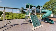 This outdoor community space is designed to offer a variety of recreational amenities for residents of all ages. At the heart of the playground area stands a multi-level play structure finished in green and white, featuring a rock climbing wall, a tire swing, traditional chain swings, a covered upper deck with safety railings, a connecting bridge, and a spiral slide. The structure is set on impact-absorbing rubberized surfacing, ensuring a safe play environment. Lush green trees and open skies frame the area, creating a vibrant and inviting atmosphere.

Adjacent to the playground, a spacious outdoor swimming pool serves as the community's central gathering point. The pool deck is finished in smooth concrete and is generously lined with lounge chairs and shaded seating areas featuring umbrellas, perfect for relaxing on sunny days. A covered porch with a ceiling fan and stone-accented columns provides a comfortable shaded retreat directly overlooking the pool.

The surrounding grounds include well-maintained grassy lawns with outdoor picnic tables and grill stations, ideal for casual gatherings. Residential buildings with classic siding, shuttered windows, and private balconies border the green space, contributing to the warm, community-oriented character of this outdoor amenity area.