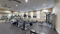 This fitness center is a comprehensively outfitted facility designed to support a full range of workout routines. The space features a durable dark rubber flooring throughout, providing both safety and comfort underfoot. The room is finished with warm beige walls, a drop-tile ceiling with recessed lighting, and multiple ceiling fans that ensure consistent air circulation throughout the space.

The cardio section is prominently featured, housing multiple treadmills, elliptical cross-trainers, and a stair climber, all from reputable brands including Precor. Large wall-mounted mirrors along one side of the room amplify the sense of space and allow users to monitor their form during workouts. Mounted flat-screen televisions are positioned for easy viewing from the cardio equipment area.

The strength training section includes a cable machine, a multi-function weight machine, a free-weight rack with dumbbells, and an adjustable workout bench, providing options for a complete resistance training session. Built-in wooden storage cubbies and wall hooks offer convenient storage for personal belongings.

Natural light filters in through multiple windows fitted with white blinds, and framed artwork adds a polished, refined ambiance to the space. A dedicated entry vestibule with glass doors provides easy access, completing this well-appointed and functional fitness facility.