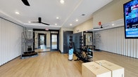 This dedicated fitness and yoga room is designed to support a wide range of workout activities in a clean, functional environment. The space features light hardwood flooring that provides a warm, inviting foundation ideal for both yoga practice and strength training. Recessed lighting throughout the ceiling ensures the room is well-lit, while ceiling fans promote comfortable airflow during active sessions. Large mirrors are strategically positioned along the walls, enhancing the sense of space and allowing for proper form checks during exercise. The room is outfitted with a variety of fitness equipment, including a medicine ball rack, kettlebells, plyo boxes, and additional strength training accessories, making it suitable for diverse workout routines. Dark-framed glass doors and entryways add a modern, polished aesthetic to the space, while neutral wall tones keep the atmosphere calm and focused. A fire alarm system and exit signage reflect the room's attention to safety standards. The overall design balances practicality with a contemporary style, creating a welcoming environment for users of all fitness levels to engage in yoga, functional training, or general exercise.