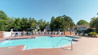 This outdoor pool area offers a welcoming resort-style retreat designed for relaxation and recreation. At its center sits a generously sized, freeform swimming pool with crystal-clear turquoise water, accessible via stainless steel entry rails and featuring a shallow wading section alongside deeper areas. The pool is complemented by an attached spa, providing an added layer of leisure.

Surrounding the pool, a expansive brick-paved sundeck provides ample space for lounging, furnished with rows of chairs and chaise lounges shaded by navy blue market umbrellas. Decorative brick columns punctuate the deck perimeter, adding a classic architectural character to the space.

The area is enclosed by a crisp white fence, lending a sense of privacy and definition to the space. Mature trees and lush greenery frame the surroundings, offering natural shade and a tranquil atmosphere. In the background, well-maintained residential buildings are visible, integrating the pool area naturally within its community setting.

Under open blue skies, the space is bathed in abundant natural light, creating a bright and inviting ambiance. Whether used for active swimming or leisurely sunbathing, this pool area is thoughtfully designed to serve as a central outdoor gathering space.
