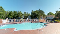 This outdoor pool area offers a welcoming resort-style retreat designed for relaxation and recreation. At its center sits a generously sized, freeform swimming pool with crystal-clear turquoise water, accessible via stainless steel entry rails and featuring a shallow wading section alongside deeper areas. The pool is complemented by an attached spa, providing an added layer of leisure.

Surrounding the pool, a expansive brick-paved sundeck provides ample space for lounging, furnished with rows of chairs and chaise lounges shaded by navy blue market umbrellas. Decorative brick columns punctuate the deck perimeter, adding a classic architectural character to the space.

The area is enclosed by a crisp white fence, lending a sense of privacy and definition to the space. Mature trees and lush greenery frame the surroundings, offering natural shade and a tranquil atmosphere. In the background, well-maintained residential buildings are visible, integrating the pool area naturally within its community setting.

Under open blue skies, the space is bathed in abundant natural light, creating a bright and inviting ambiance. Whether used for active swimming or leisurely sunbathing, this pool area is thoughtfully designed to serve as a central outdoor gathering space.