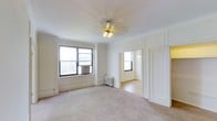 This single-floor space showcases classic architectural details that lend it a timeless character. The main living area is finished with soft neutral carpeting and crisp white walls adorned with elegant panel moldings, contributing to a clean and refined aesthetic. A gold-toned ceiling fan light fixture serves as a subtle decorative focal point overhead, complementing the traditional styling of the room.

Natural light enters generously through black-framed windows, one of which houses a wall-mounted air conditioning unit, while a cast-iron radiator beneath provides heating — a hallmark of classic building construction. The white walls and light-toned flooring work together to enhance the sense of brightness and openness throughout.

An open doorway leads into an adjoining room, also flooded with natural light from its own window, suggesting a well-connected and functional layout. The consistent use of white paint, panel detailing, and period-appropriate fixtures across both visible spaces creates a cohesive and airy atmosphere. Overall, the space blends classic architectural character with a neutral, versatile interior palette.