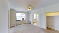 This single-floor space showcases classic architectural details that lend it a timeless character. The main living area is finished with soft neutral carpeting and crisp white walls adorned with elegant panel moldings, contributing to a clean and refined aesthetic. A gold-toned ceiling fan light fixture serves as a subtle decorative focal point overhead, complementing the traditional styling of the room.

Natural light enters generously through black-framed windows, one of which houses a wall-mounted air conditioning unit, while a cast-iron radiator beneath provides heating — a hallmark of classic building construction. The white walls and light-toned flooring work together to enhance the sense of brightness and openness throughout.

An open doorway leads into an adjoining room, also flooded with natural light from its own window, suggesting a well-connected and functional layout. The consistent use of white paint, panel detailing, and period-appropriate fixtures across both visible spaces creates a cohesive and airy atmosphere. Overall, the space blends classic architectural character with a neutral, versatile interior palette.