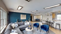 This single-floor community space is thoughtfully designed with a cohesive modern aesthetic, blending bold color accents with neutral tones throughout. The main lounge area serves as the centerpiece, anchored by a large sectional sofa in charcoal gray, complemented by plush royal blue accent chairs and a patterned area rug. A striking teal feature wall adorned with large-scale artwork adds visual depth and character to the space. Flush-mounted rectangular light fixtures provide consistent, well-distributed illumination across the ceiling.

Large windows along one side of the lounge frame views of an outdoor pool area, filling the space with natural light and creating a seamless connection to the exterior. A wall-mounted flat-screen television and a decorative console with mirrored paneling enhance the entertainment-oriented atmosphere.

Adjacent to the lounge is a dedicated dining and workspace area, outfitted with bar-height seating and a compact workstation. A hallway lined with tile flooring leads deeper into the space, connecting to a functional kitchenette. The kitchen features light wood cabinetry, white countertops, stainless steel appliances, and a herringbone mosaic tile backsplash, combining practicality with polished design details throughout.