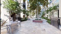 Located at 2900 West Dallas Street in Houston's prestigious River Oaks corridor, this outdoor amenity space is a beautifully designed courtyard retreat set within a multi-story residential complex. The centerpiece of the space is a generously sized resort-style swimming pool with clear, inviting blue water, complemented by a shallow wading area with in-pool lounge chairs — ideal for relaxing in the Houston sun. The pool deck is finished with elegant natural stone pavers that extend throughout the courtyard, providing a clean and sophisticated aesthetic. Multiple chaise lounges are thoughtfully arranged along the poolside, offering ample space for sunbathing and leisure. Raised brick planters anchor the landscape design, filled with mature trees including towering palms and broad-canopy shade trees that lend a lush, resort-like atmosphere to the enclosed courtyard. Well-maintained greenery and manicured shrubs line the building perimeters, softening the architecture and adding natural vibrancy. The surrounding multi-story building facades feature neutral tones, large windows, and private balconies that overlook the courtyard below. Additional seating areas with outdoor dining furniture are positioned throughout the space, making it suitable for both casual gatherings and quiet relaxation. The overall ambiance is serene, polished, and resort-inspired.