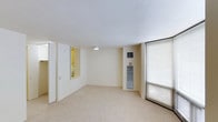 This single-floor space presents a clean, minimalist design centered around a neutral color palette of soft whites and warm beige tones. The main living area is finished with plush wall-to-wall carpeting underfoot, contributing to a quiet and comfortable atmosphere throughout. A large window fitted with horizontal blinds dominates one wall, allowing natural light to filter into the room while providing privacy and light control. The textured ceiling adds subtle architectural character to the otherwise streamlined interior. A pass-through window or interior opening connects the main area to an adjacent room, which appears to be illuminated and lined with shelving or cabinetry, suggesting a functional storage or utility space. Along one wall, a slim vertical unit — likely a wine cooler or narrow storage appliance — sits beneath a wall-mounted control panel, adding a practical element to the space. The overall layout is open and uncluttered, offering flexibility for various furnishing arrangements. The space maintains a consistent, understated aesthetic that lends itself well to personalization, making it a versatile and functional environment suited for a variety of uses.