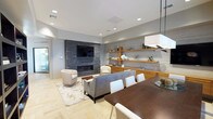 This single-floor space presents a cohesive, contemporary design that seamlessly integrates multiple functional areas. The open-concept layout connects a living room, dining area, and lounge, creating a fluid and spacious environment well-suited for both relaxation and gatherings.

The living area is anchored by a dramatic floor-to-ceiling stacked stone fireplace wall, which houses a mounted flat-screen television and serves as a striking focal point. Floating wood shelves and a low-profile wood media credenza line an adjacent textured wall, offering both display and storage functionality. Plush gray seating, a geometric-patterned area rug, and a warm wood coffee table complete the lounge setting.

The dining area features a substantial dark wood table surrounded by upholstered chairs, illuminated by a rectangular pendant light fixture. Floor-to-ceiling drapery frames large windows, allowing natural light to fill the space. A floor-to-ceiling dark wood bookcase adds depth and visual interest nearby.

Throughout the space, light herringbone-patterned hardwood flooring provides a unifying element. Recessed lighting and tray ceiling details contribute to the refined, polished ambiance. A connecting corridor with exposed beam accents offers glimpses into additional adjoining areas, reinforcing the open and well-designed character of this space.