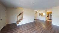 This two-story residence is designed with a practical and straightforward layout. The main level features a spacious living area finished with rich, dark hardwood flooring that extends throughout, complemented by neutral cream-toned walls. A carpeted staircase with a wooden banister connects to the upper level. The kitchen is efficiently arranged with warm oak cabinetry, granite-style countertops, a tile backsplash, and stainless steel appliances including an electric range and dishwasher. A window above the sink provides natural light and an outdoor view. Two half-bathrooms are present, each featuring tile-style flooring; one includes a gray shaker-style vanity with a white countertop, while the other is more utilitarian with grab bars and baseboard heating. The upper floor hosts multiple carpeted bedrooms bathed in natural light from double windows. Baseboard heating is a consistent feature throughout the bedrooms. Closet space is provided in the bedrooms, including a dedicated walk-in style closet. A utility area houses the water heater and associated mechanical systems. Overall, the space presents a clean, functional aesthetic suited for comfortable everyday living.