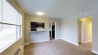 This single-floor unit offers a clean, functional layout designed to maximize its use of space. The living area features neutral-toned wall-to-wall carpeting and light gray walls, creating a simple, understated atmosphere. Natural light filters in through a window along one side, brightening the interior. A pass-through cutout connects the living area to the kitchen, enhancing the sense of openness between the two spaces. The kitchen is outfitted with dark espresso-toned cabinetry, a light-colored countertop that doubles as a breakfast bar, and black appliances including a microwave and refrigerator. A doorway from the living area leads further into the unit, providing access to additional spaces. Flush-mount ceiling lighting and clean white baseboards add to the tidy, well-maintained appearance throughout. The overall ambiance is practical and move-in ready, suited for those seeking a straightforward, low-maintenance living space.