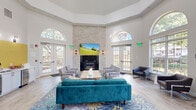 Located at 100 Hunsberger Dr in Limerick, PA, this single-floor living room is a thoughtfully designed communal space that combines architectural elegance with comfortable, modern furnishings. The room's most striking feature is its dramatic ceiling height, highlighted by a coffered tray ceiling that adds depth and a sense of grandeur. Multiple large arched windows flood the interior with abundant natural light and offer views of the surrounding greenery, creating a warm and inviting atmosphere throughout the day. At the heart of the room stands a floor-to-ceiling stacked-stone accent wall, anchored by a sleek fireplace below and a wall-mounted flat-screen television above, serving as a sophisticated focal point. The seating arrangement is both functional and stylish, featuring a vibrant teal velvet sofa, coordinating gray accent chairs, and a light wood coffee table, all set atop a bold floral-patterned area rug laid over light hardwood flooring. A compact kitchenette with a yellow tile backsplash, stainless steel appliances, and white cabinetry adds practical convenience. Decorative wall art, wall sconces, and French doors leading to the outdoors complete the polished, welcoming character of this well-appointed space.
